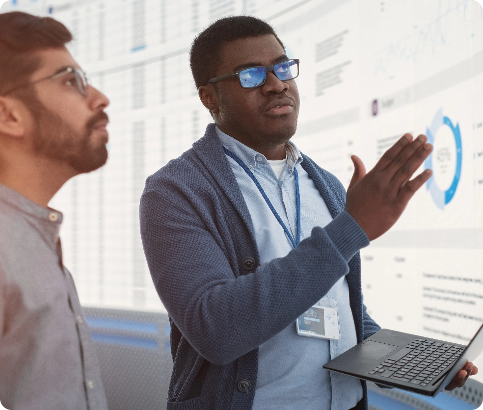 Businessman wearing glasses holds a laptop and gestures at a large screen full of data while brainstorming with a coworker 
