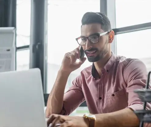 Man with glasses and dark hair on phone looking at a computer screen