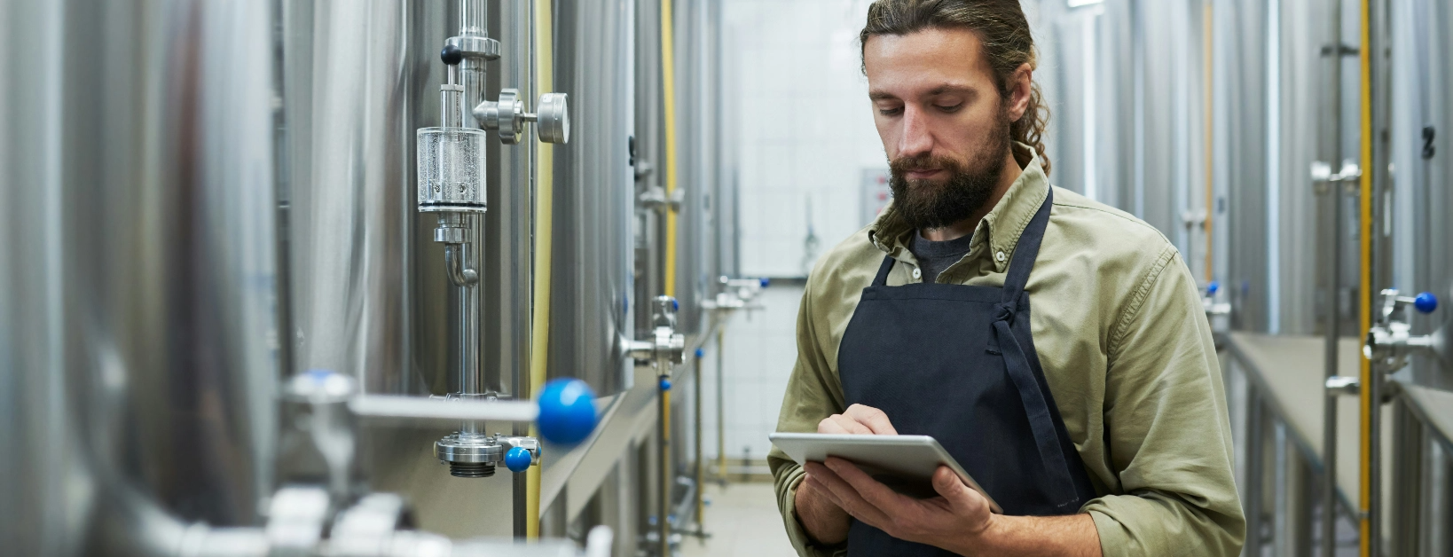 Bearded man wearing an apron uses brewery and distillery ERP on a tablet with brewing equipment in the background
