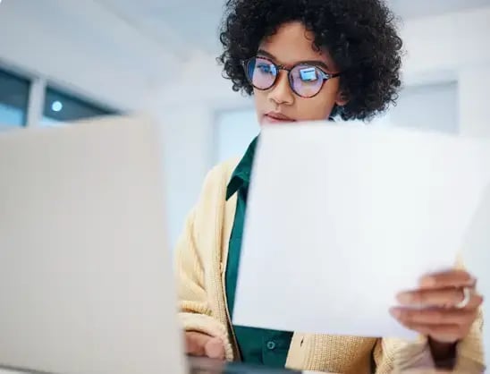 Businesswoman wearing glasses compares intercompany accounting data on a piece of paper to the data on her laptop