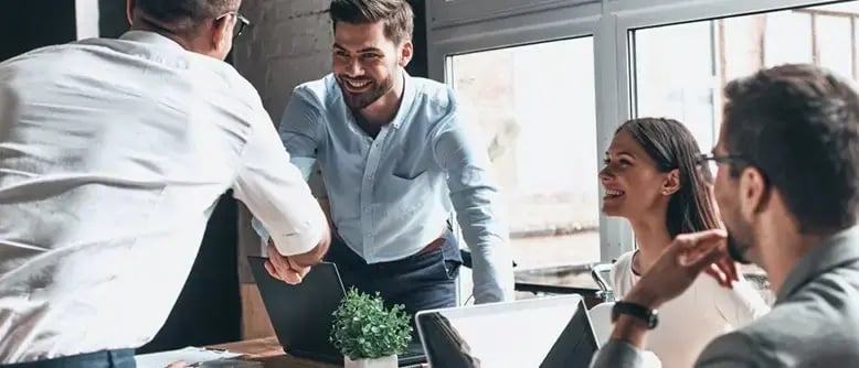 Businessman and businesswoman smile at two businessmen shaking hands at a meeting