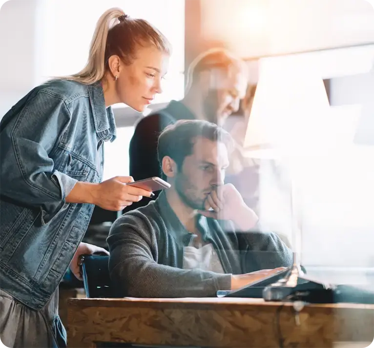 Two businesspeople collaborate at a desk, looking at a computer screen, with a person in the background
